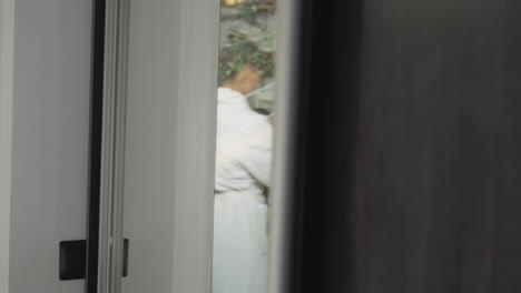 woman cooks in kitchen view through glass door. lady in white bathrobe prepares breakfast standing by countertop at home. domestic lifestyle scene