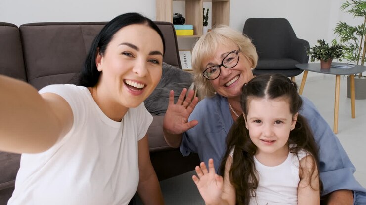Nonna, madre e bambina che salutano e guardano la telecamera mentre fanno una videochiamata tramite tablet sedute sul pavimento a casa.