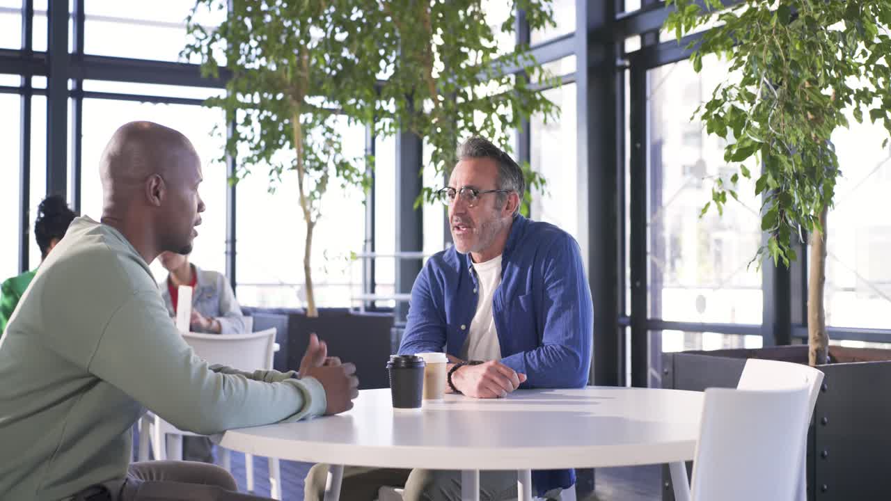 After grabbing mugs, two male colleagues walking to lounge table, discussing project, shaking hands