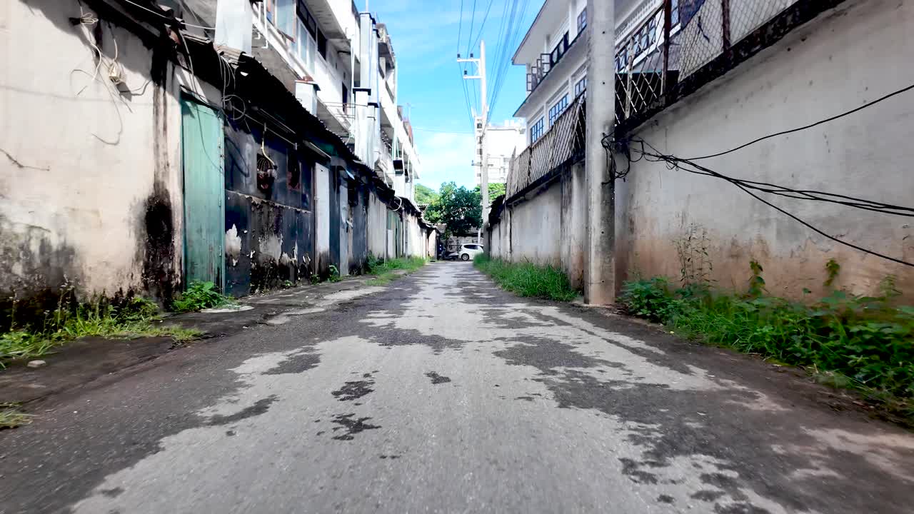 Urban residential alleyway with buildings, walls, and overgrown weeds under a clear sky