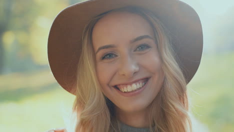 vista de cerca de una joven rubia con sombrero y sonriendo a la cámara en un parque por la mañana