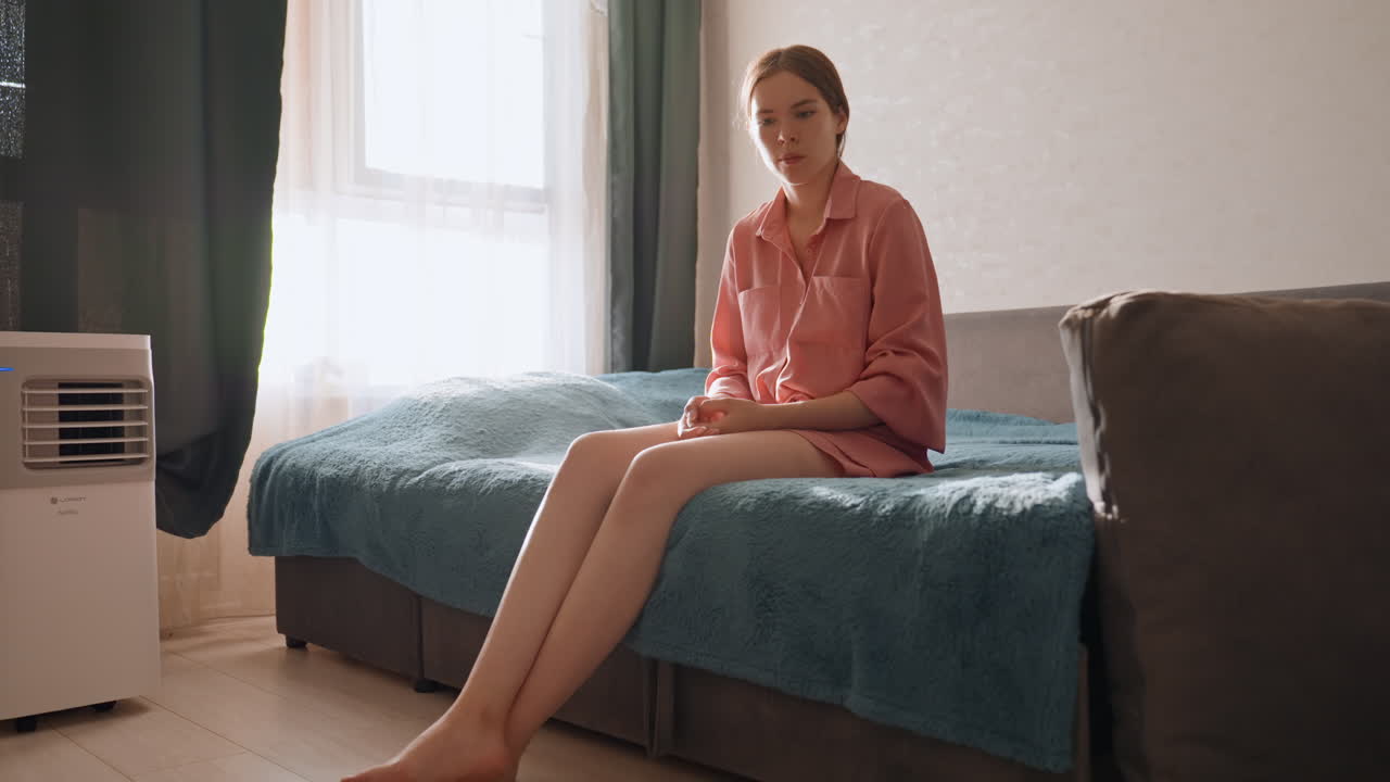 Caucasian Woman Sitting On Bed, Soft Morning Light Through Window, Teal Blanket, Small Fluffy Puppy At Feet, Pensive Expression, Quiet Apartment Interior, Intimate Domestic Mood, Slow Reflective