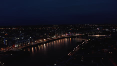 Vista-Panorámica-Aérea-Del-Puente-Iluminado-Y-El-Paseo-Marítimo-De-La-Ciudad.-Luces-Que-Reflejan-La-Superficie-Del-Agua.-Río-Shannon-Que-Fluye-Tranquilamente-En-La-Ciudad-Nocturna.-Limerick,-Irlanda