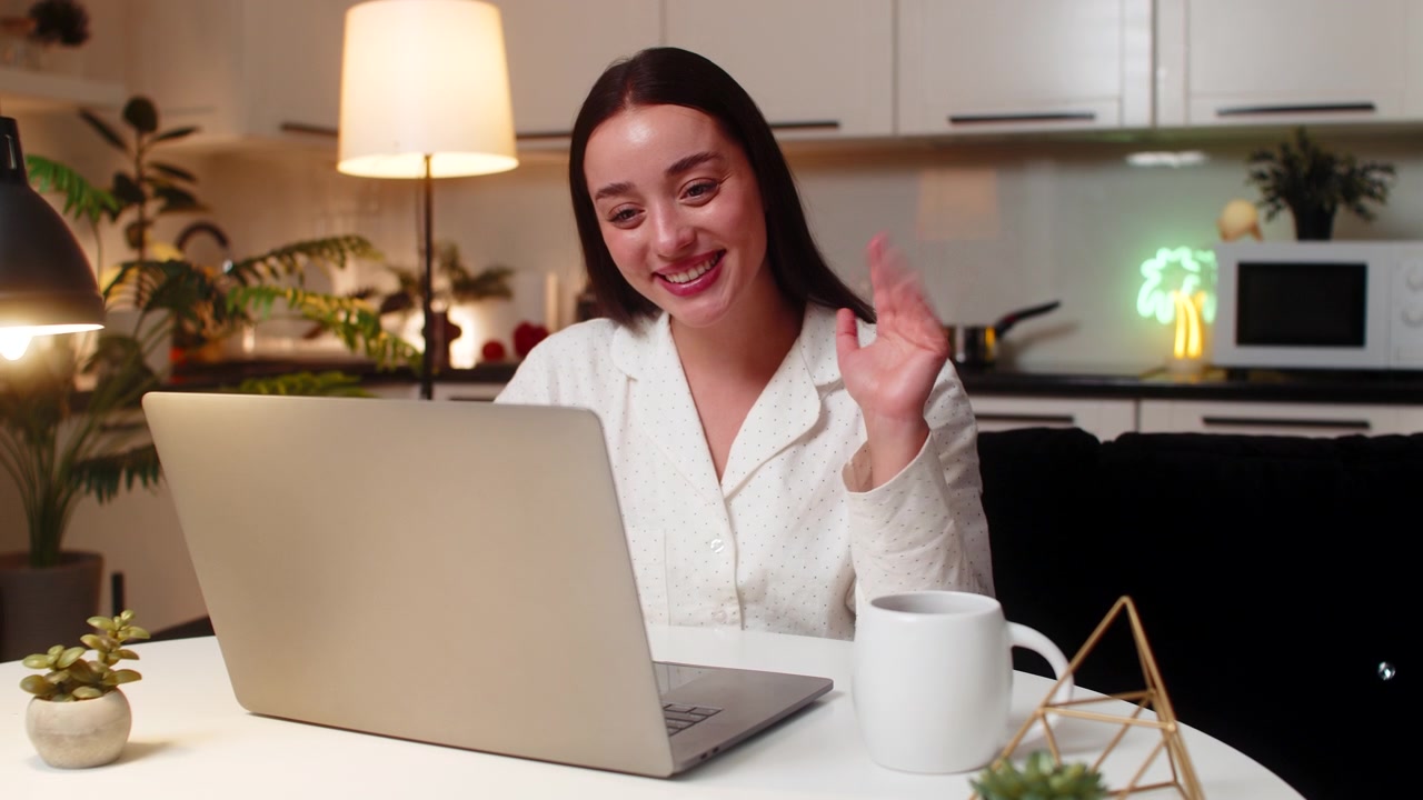 Mujer joven saluda por la webcam de su portátil, habla, muestra gráficos al cliente, escucha y asiente en una reunión online por la noche