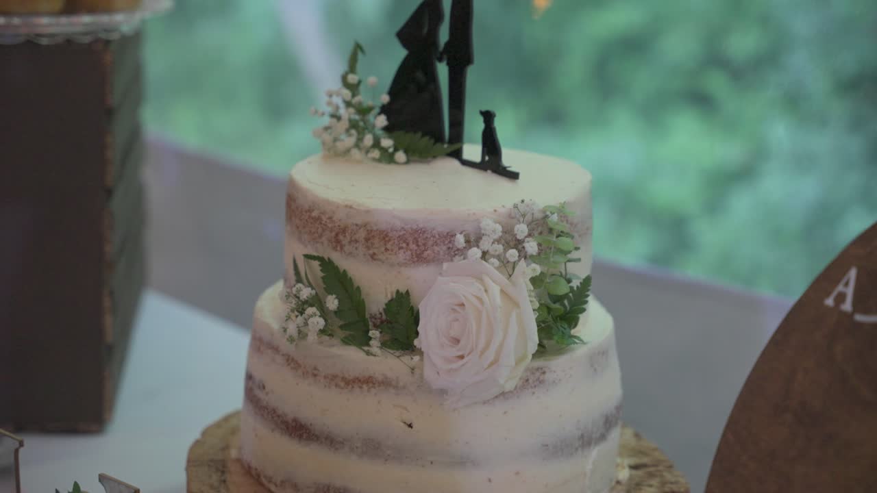 Uma inclinação suave para cima de um bolo de casamento branco adornado com uma rosa fresca e folhagem, apresentando um topo de bolo com silhuetas de noiva, noivo e cachorro sobre uma base de madeira natural.