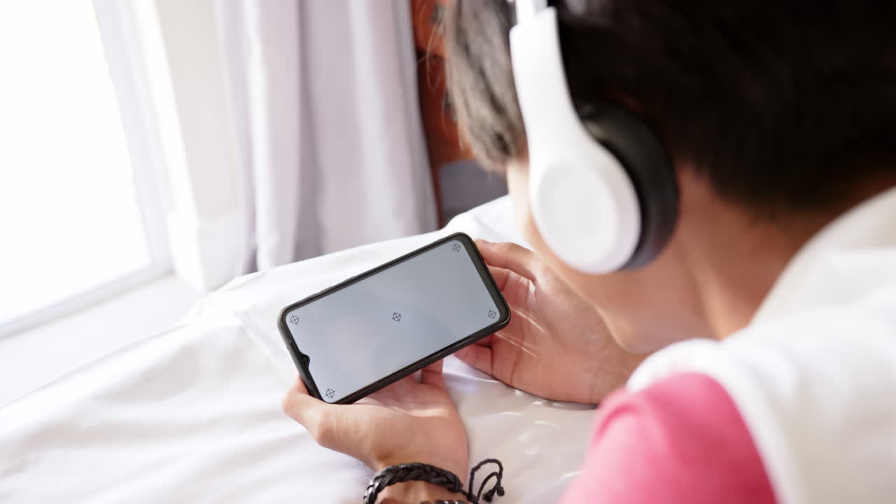 Wearing headphones, person watching video on smartphone while lying on bed