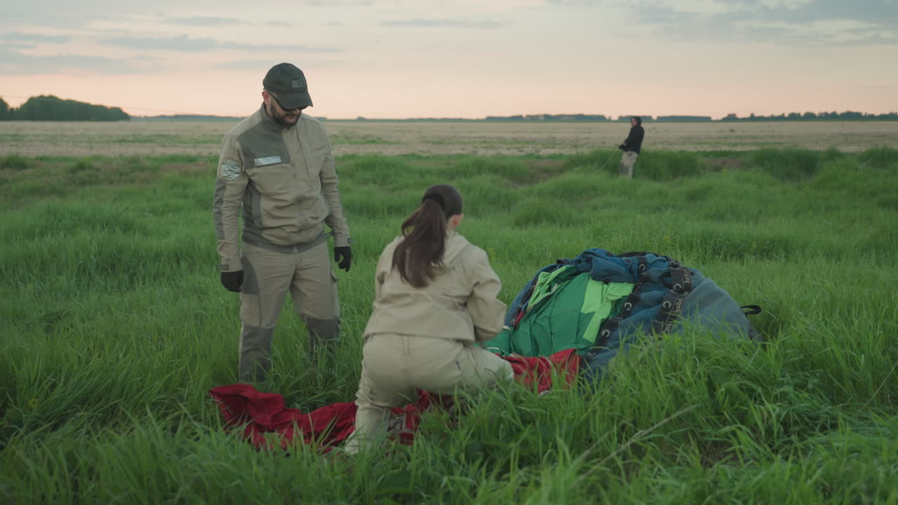 man in cap jacket watches woman in pale jacket calmly pulling red balloon envelope cloth from duffel bag on grassy field during preflight setup with another crew member in background