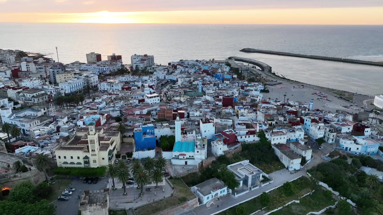 Vista aérea amplia de Asilah, Marruecos, mostrando densos barrios blancos y coloridos que se extienden hacia la costa atlántica, con la histórica ciudad costera y el mar de fondo.