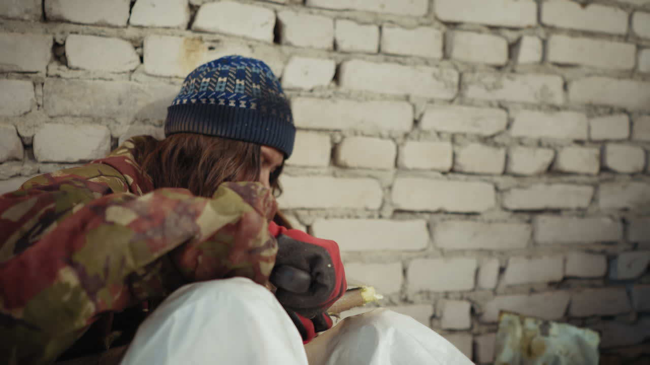 겨울 모자와 빨간 장갑을 낀 사냥꾼이 벽돌담 옆에 앉아 사냥용 칼로 긴 막대기를 날카롭게 갈고 있어요. 추운 버려진 방에서 거친 위장 재킷을 입고 생존을 위해 집중하는 모습이에요.