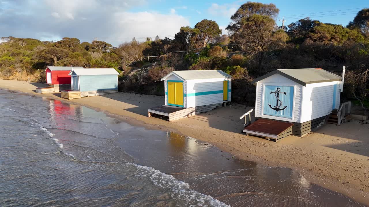 Drone glides above sandy beach, vibrant bathing boxes, gentle waves, and coastal vegetation in sunlight