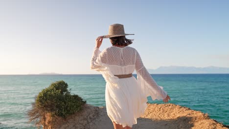 young fashionista woman on cliff over ocean with teal water. lady in airy dress and hat exploring island nature. she spending holiday trip at amazing landmark - luxury resort. golden hour.