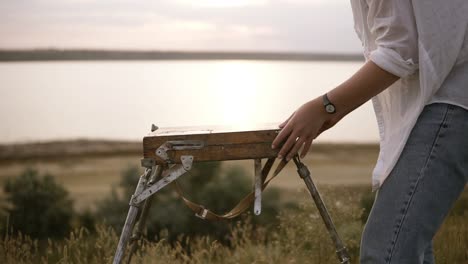 young caucasian brunette ispreparing her easelfor work outdoors. clear meadow with the blurred view of lake on the background