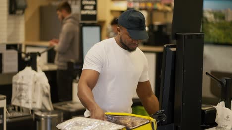 a delivery man with black skin in a white t-shirt and a black cap puts all the necessary goods after buying them into a large yellow sealed bag in order to deliver them. male delivery man purchases necessary goods in the store