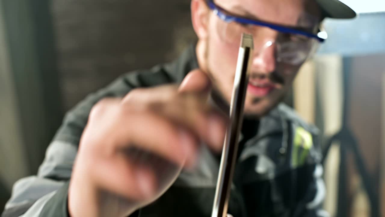 Konzentrierter junger bärtiger Schreiner mit Schutzbrille und Kappe prüft das Werkstück.