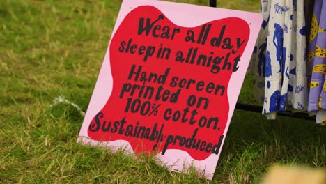 pinkish red sign with black text about sustainable produced clothing laying down on the ground in front of shop selling dresses leaning on metal hanger vegan clothes ecologically healthy recycling