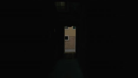 narrow, dark alleyway in venice, with a view of a brightly lit, modern residential building at the end