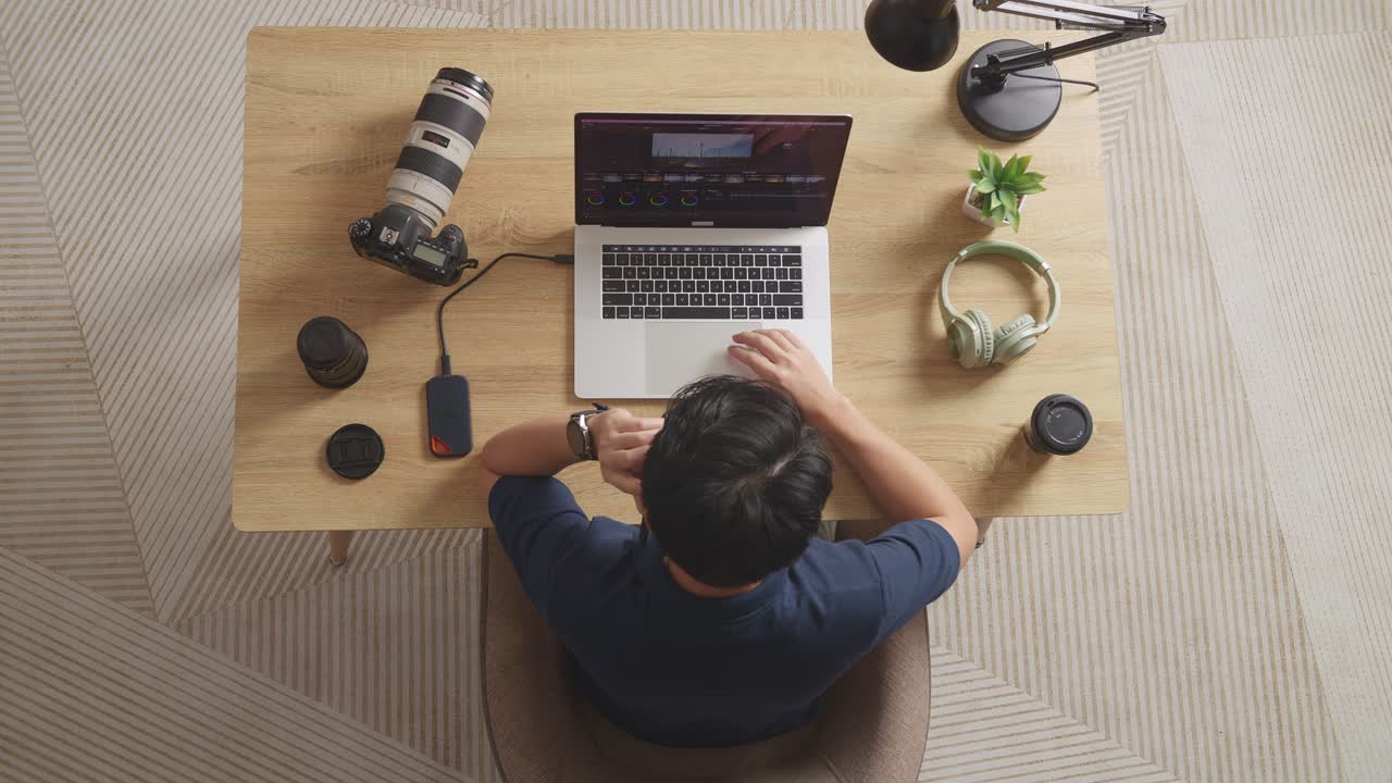 Top View Of A Male Color Grading Talking On Smartphone While Sitting In The Workspace Using A Laptop Next To The Camera Editing The Video At Home