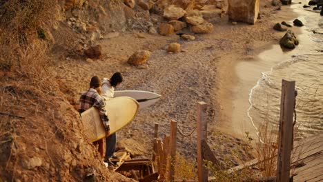 Top-view-of-a-happy-couple,-a-guy-in-a-white-sweatshirt-and-his-blonde-girlfriend-in-a-checkered-shirt-are-carrying-boards-for-skating-on-the-Surface-and-going-down-to-the-sea-near-a-rocky-shore-in-the-morning-at-Sunrise-in-fall