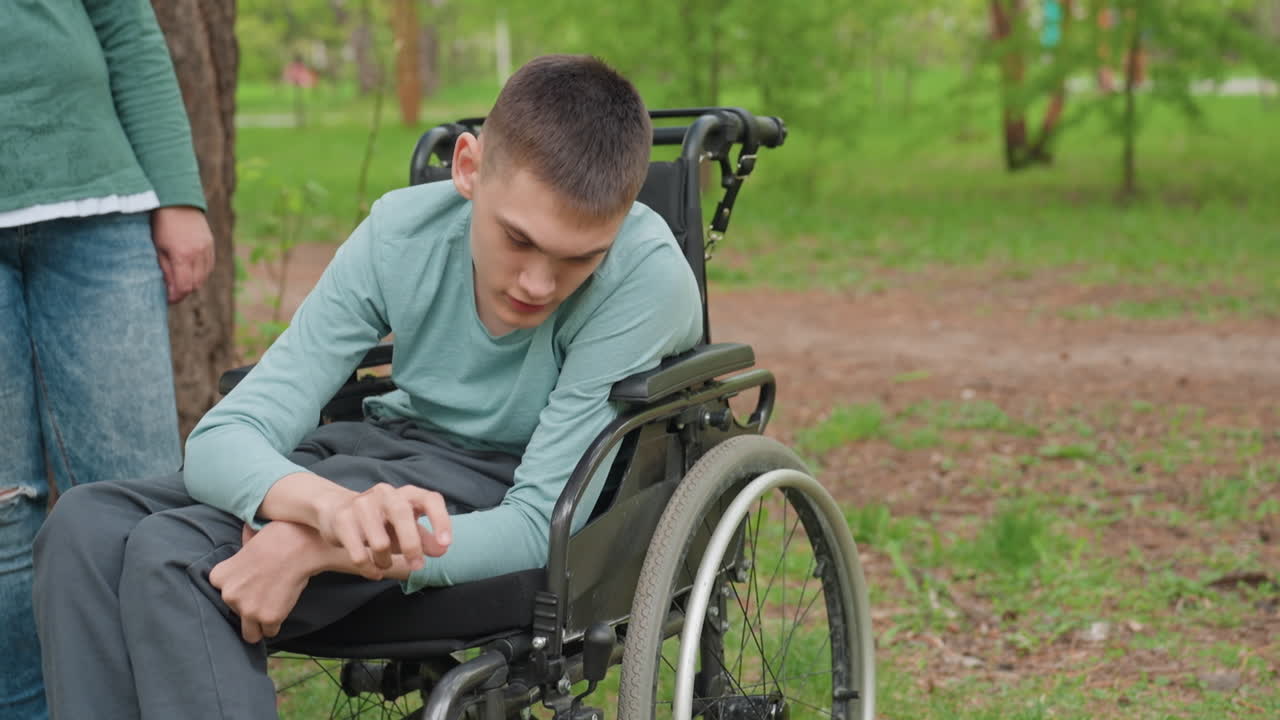 Young Man In Wheelchair In Park, Leaning Forward While Caregiver Stands Beside, Pigeons Nearby, Spring Grass And Trees, Contemplative Expression, Mobility Support And Resilience In Outdoor