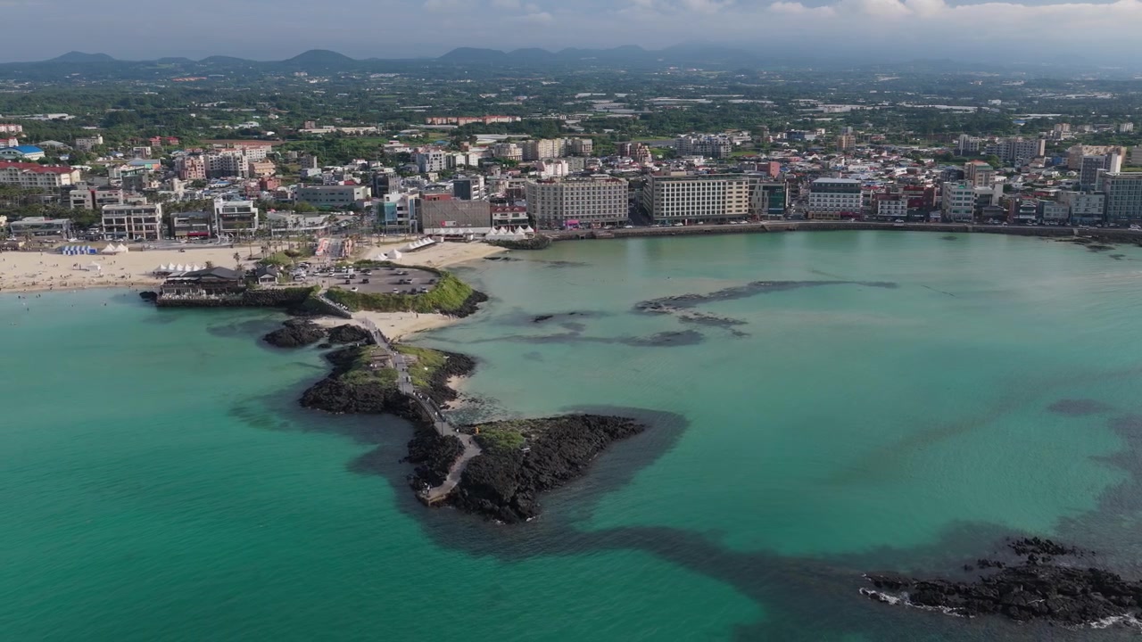 Ripresa orizzontale in cameo con carrellata all'indietro verso sinistra sopra la spiaggia di Hamdeok, Sinheung-ri, Isola di Jeju, Corea del Sud, che mostra acqua turchese, rocce vulcaniche, porto, villaggio e la costa