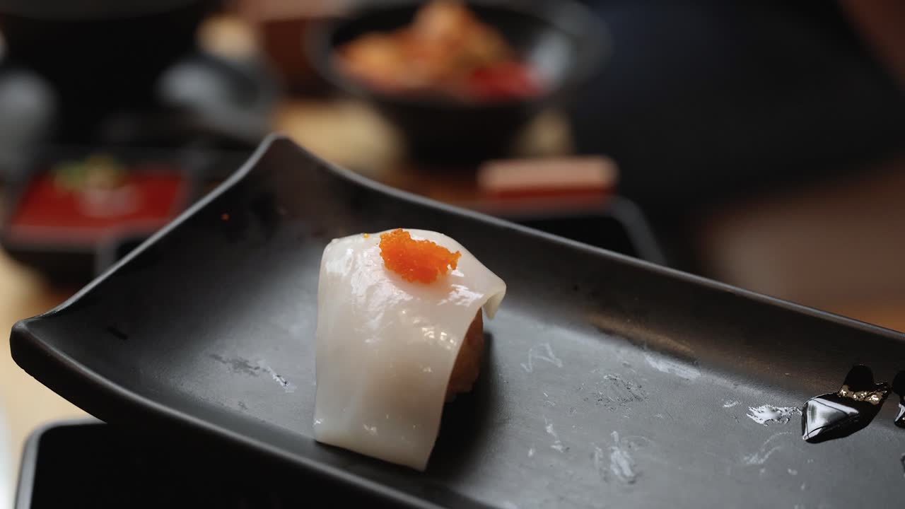 Chopsticks pick up fresh squid nigiri with tobiko from a black plate in soft lighting