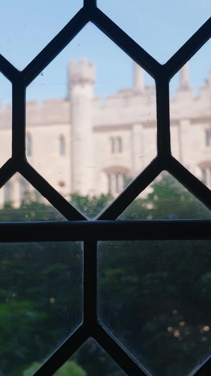 Arundel Castle courtyard seen through leaded glass window as camera pans right from inside stone room, revealing historic architecture, inner garden greenery and medieval atmosphere. Vertical.