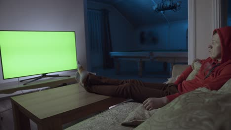 man at night sits at home on the couch, his feet on the table, remote control switches channels
