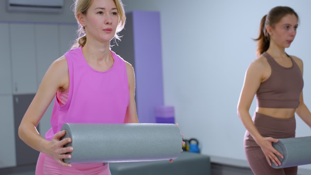 Close-up of a woman stretching forward holding a foam roller, with another woman nearby, bright light and gym equipment are visible in the background