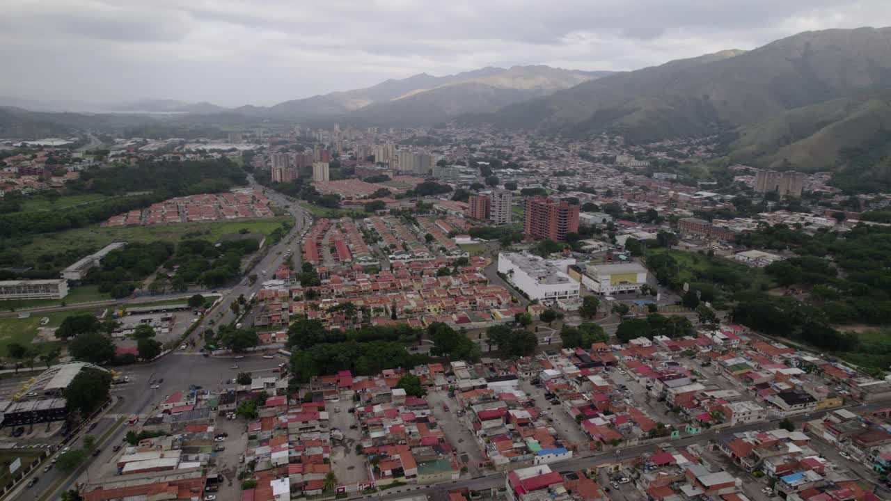 sweeping drone shot capturing Maracay, Venezuela, where modern buildings and dense housing transition into lush green foothills. Contrast development and poverty