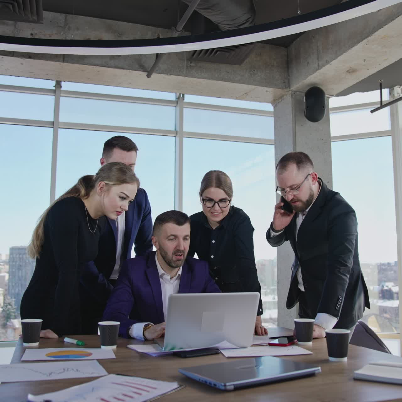 Collaborative team crowded together around their ceo in office. Man in center at computer proposes ideas to discuss. One employee answers the phone. Close up with gradual distancing