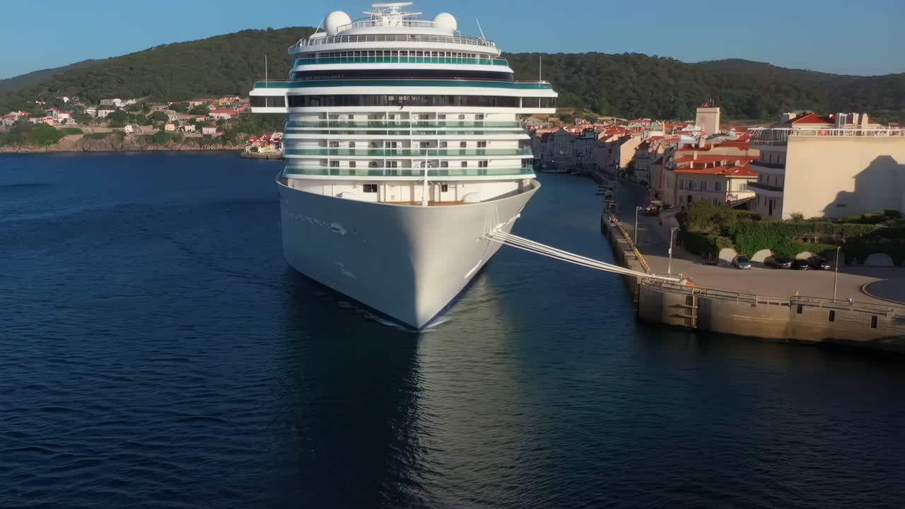 A large cruise ship navigating a coastal port with a colorful town and green hills in the background