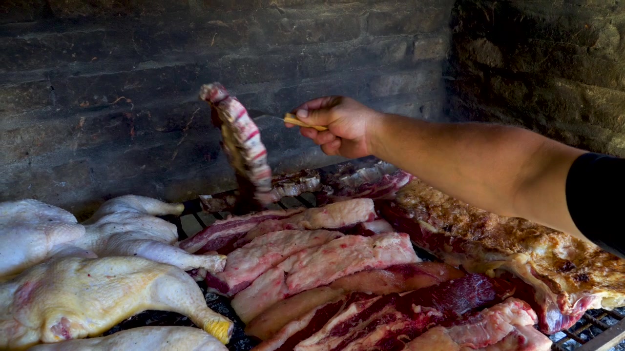 Ayam utuh dan irisan daging babi dimasak di atas panggangan bata sementara sebuah tangan meraih untuk memindahkan daging di atas panas, berguna untuk konten budaya makanan atau memasak