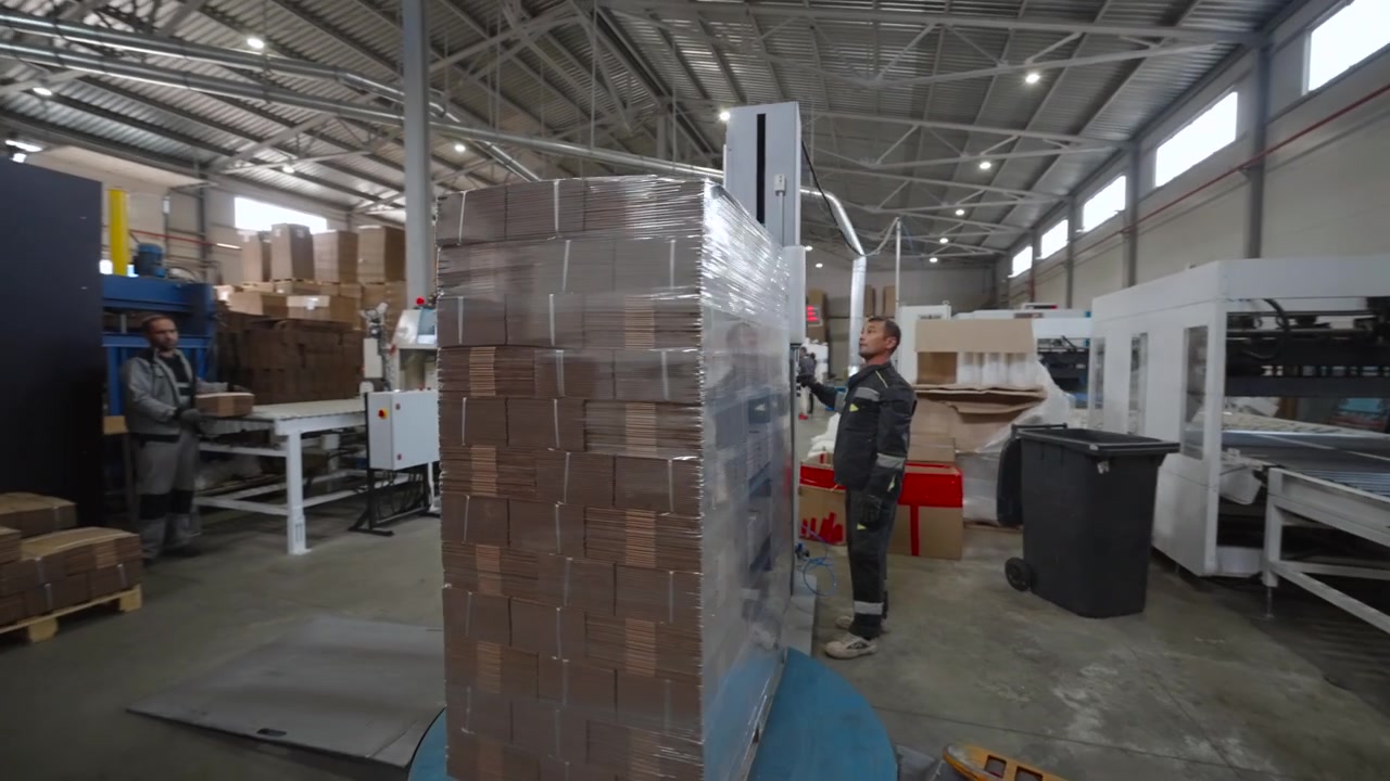 Un operaio è accanto a una macchina avvolgitrice per pallet, che fissa un'alta pila di scatole di cartone termoretraibili su un pallet di legno all'interno di un luminoso magazzino industriale con scaffalature, impianti di servizio e aperto