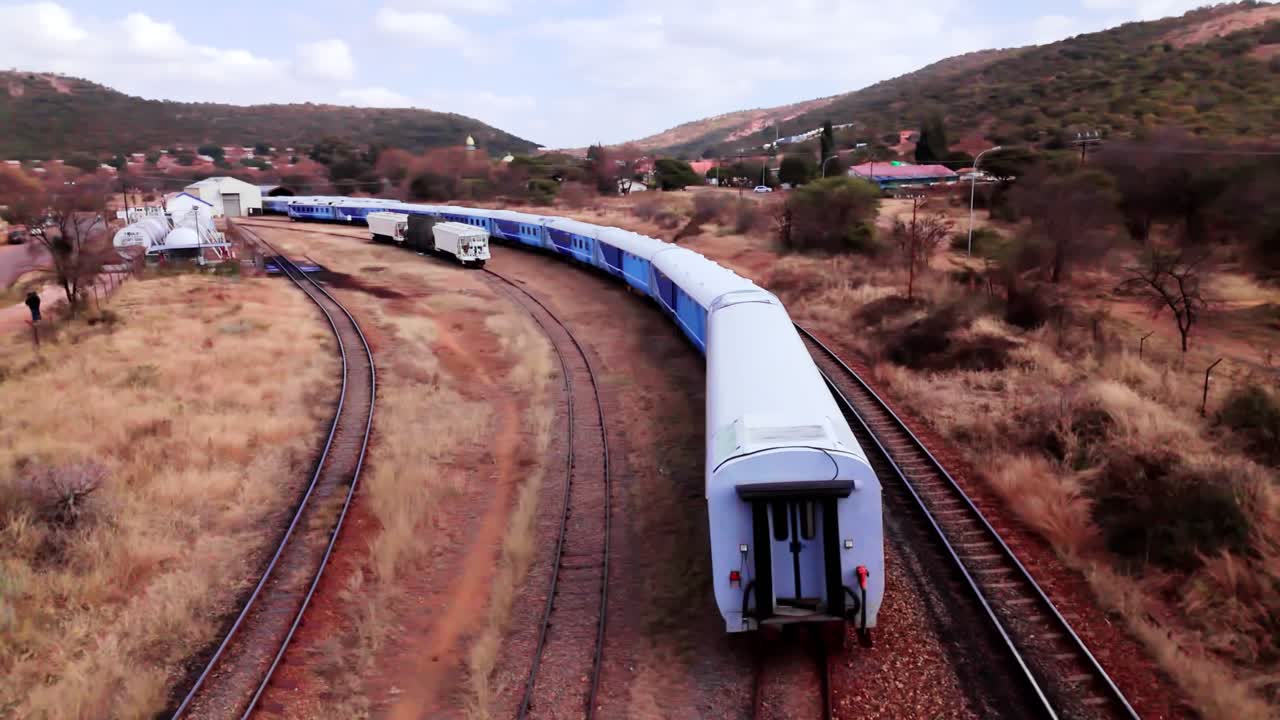 Wide Smooth Panning 4K Drone Shot - Establishing Blue Botswana Train , on a Sunny Hot Day With Dry Grass Vegetation and Brown Soil surrounding the railway line