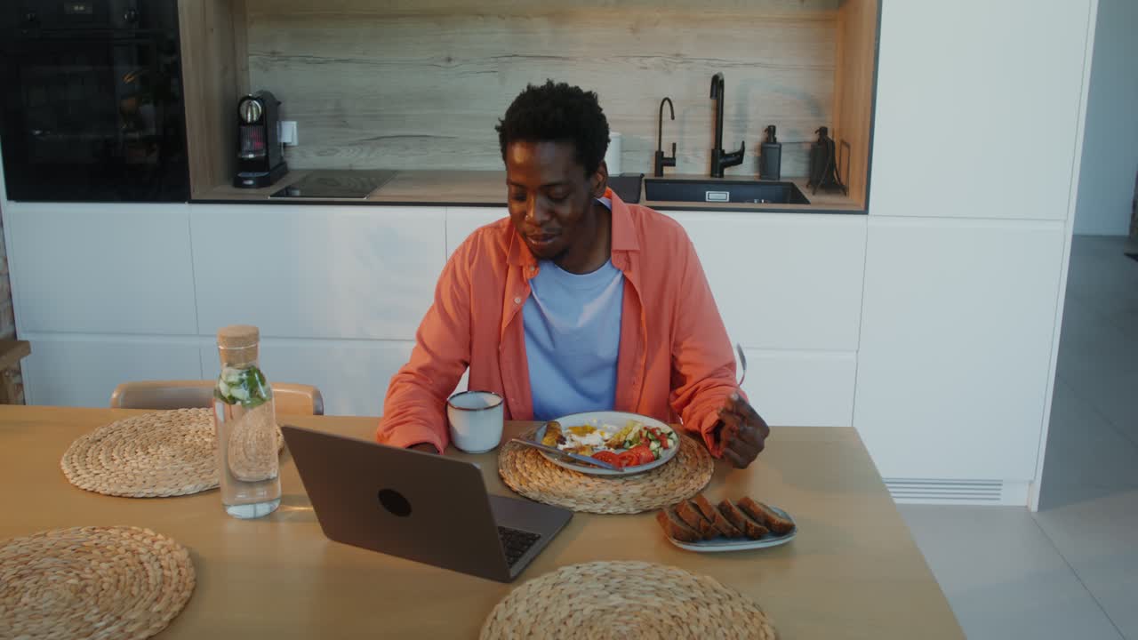 Mann spiser og jobber på laptop på kjøkkenet hjemme