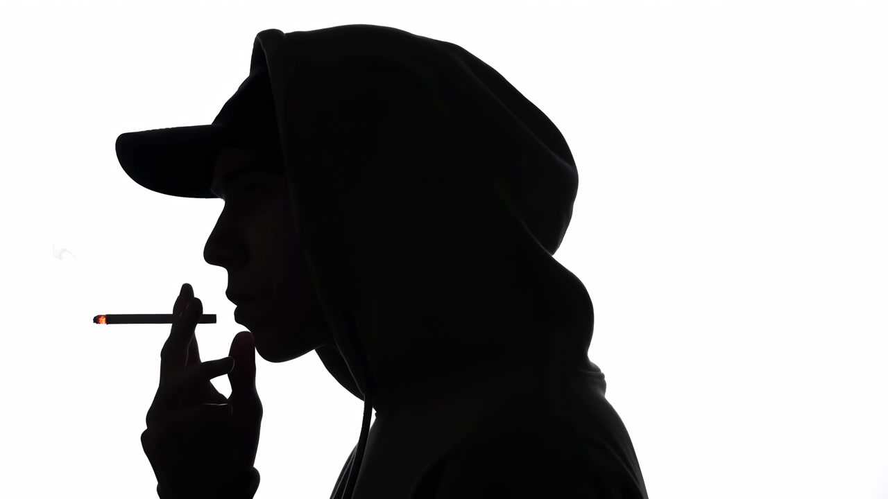 Figura scura di profilo con cappuccio e berretto tiene una sigaretta accesa vicino alle labbra mentre appare una piccola scia di fumo, creando una composizione minimalista ad alto contrasto e netta.