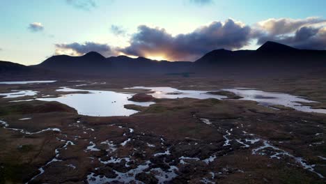 drone footage flying above of a patchwork wetland landscape of islands and peat bogs surrounded by fresh water looking towards dark mountains on the horizon at sunset