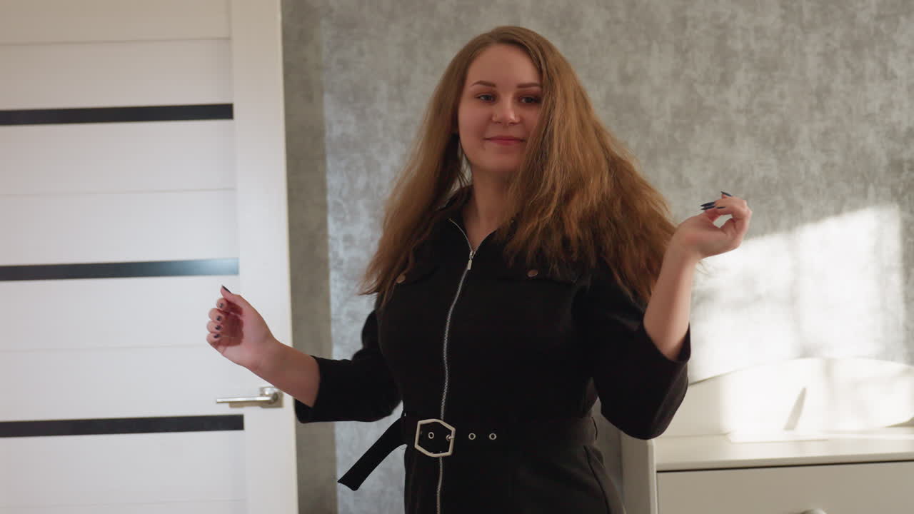 검은색 드레스를 입은 우아한 젊은 여성이 자신감 넘치는 자세로 긴 자연스러운 머리를 휘날리며 방으로 우아하게 걸어 들어가요. 부드러운 햇빛이 그녀의 매력과 균형 있는 움직임을 강조하고 있어요.