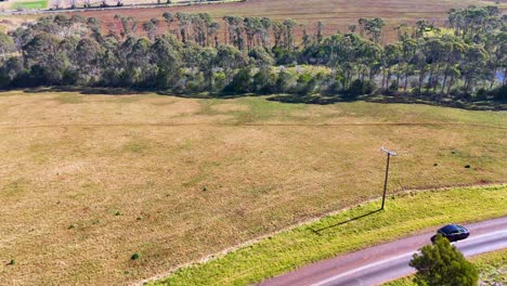 a black car travels along a winding country road bordered by grassy fields and trees in bright daylight, captured in a smooth aerial tracking shot