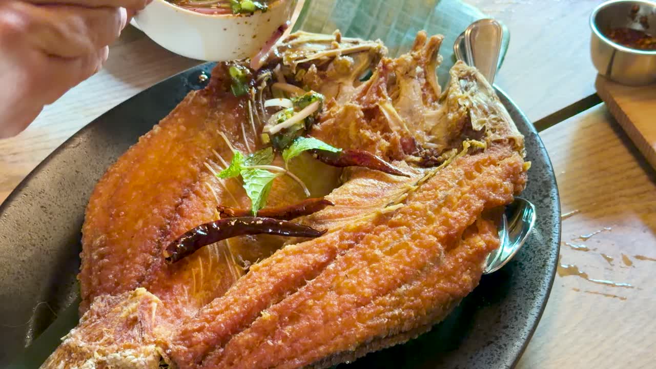 Hand pours chili herb sauce onto crispy fried fish in bright, natural light, close-up view