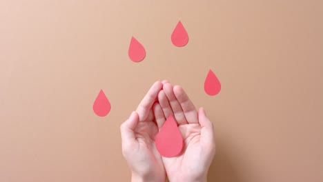 Hands-of-caucasian-woman-holding-blood-drop,-with-drops-on-beige-background,-slow-motion