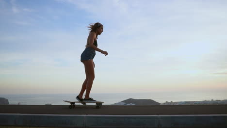 slow-motion recording captures a beautiful and stylish skateboarder in shorts riding her board along a mountain road at sunset, with the mountains creating a stunning background