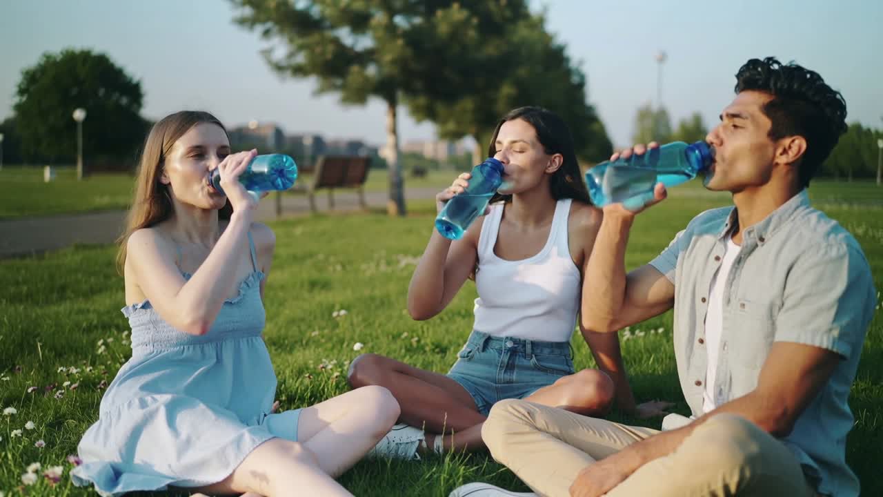 Drinking friends in summer wear hot in sun at park, talking, man gesturing, holding blue bottles