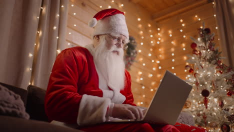 santa claus is working on the computer at home in the magical christmas lights on the background of a decorated christmas tree