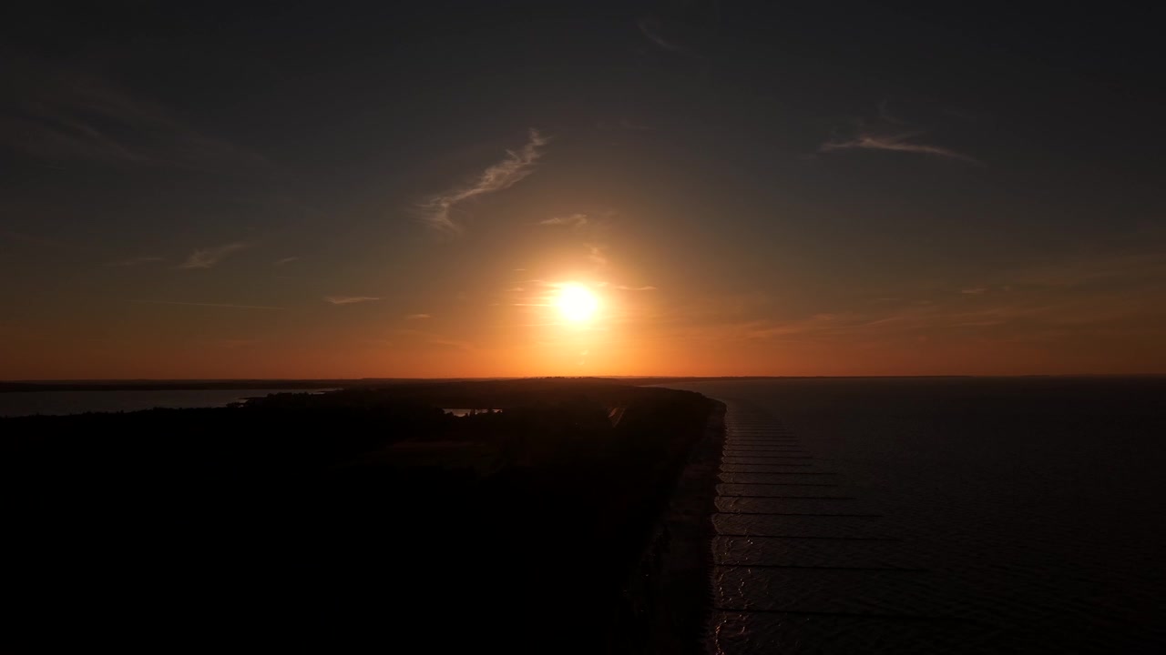 Silhuet af en træmole, der strækker sig ud i mørkt, roligt vand under en glødende gyldenorange sol, med tynde skyer, der trækker hen over den aftagende solnedgangshimmel langs kystlinjen i USA. Bredt dronebillede