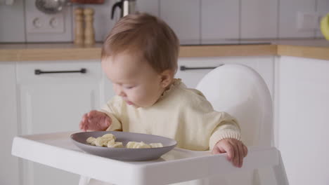 Linda-Niña-Comiendo-Rodajas-De-Plátano-Sentada-En-Su-Silla-Alta-En-La-Cocina-3