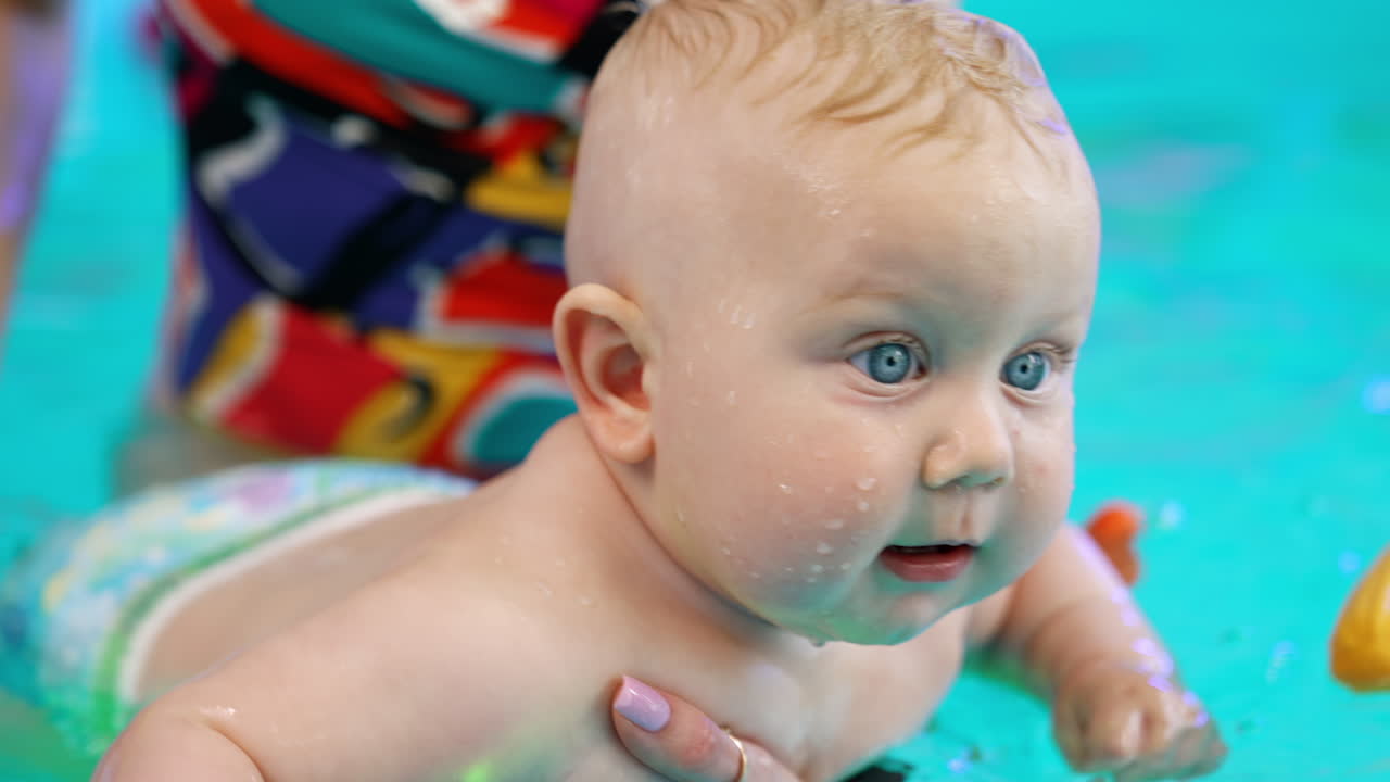 Lovely blond baby holding a toy in the water. Adorable child is learning to swim in the swimming pool. Close up.