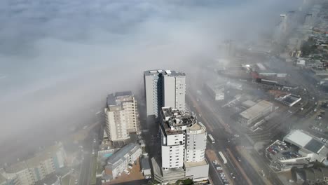 dynamic aerial view of three tall buildings in a busy coastal town with a thick layer of fog covering the ocean begind