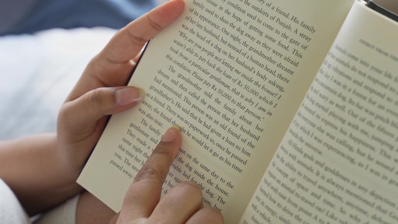 Close-up of a woman's hand gently pressing and smoothing an open book page while reading, with clearly visible printed text and soft natural window light illuminating.