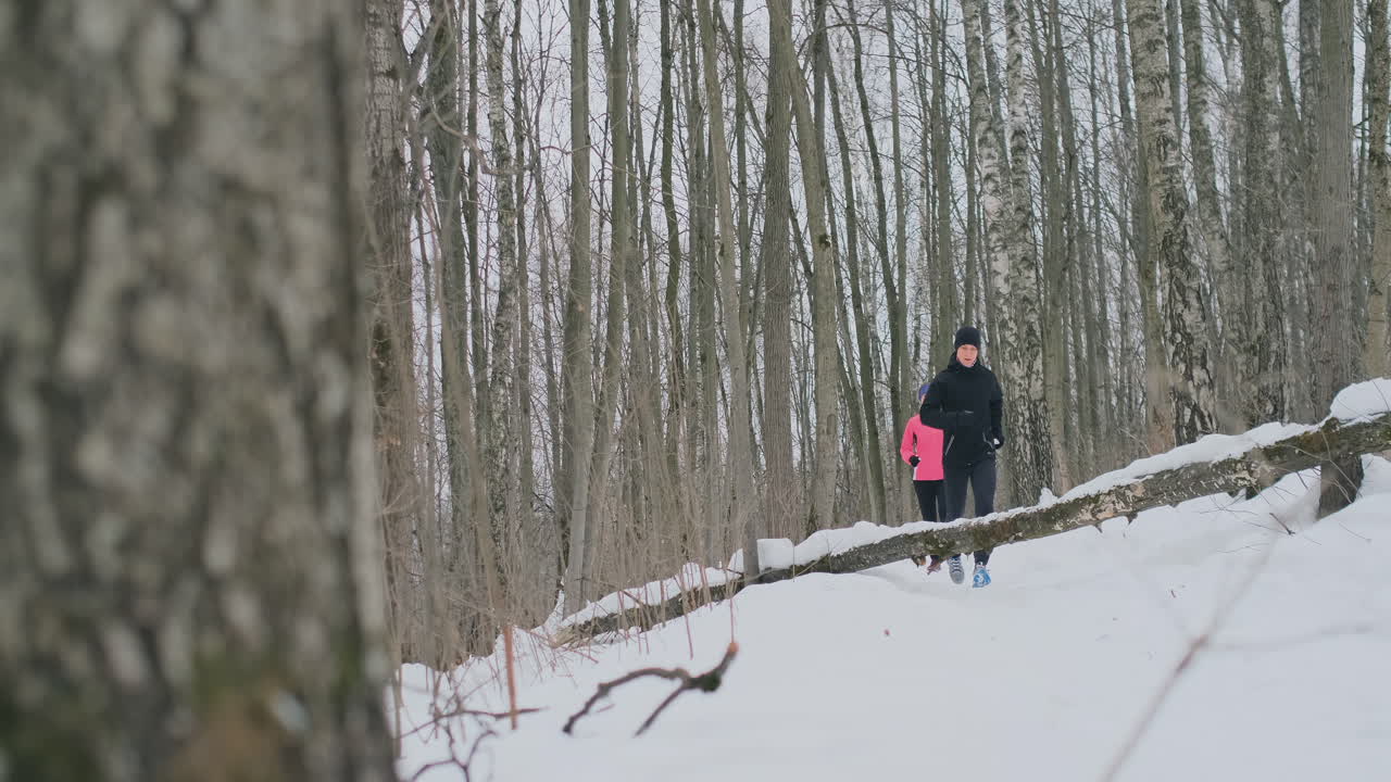 una pareja joven y saludable, positiva y hermosa, corriendo con ropa deportiva por el bosque en la soleada mañana de invierno. salta sobre el árbol, supera las dificultades del camino. pasar por encima de un obstáculo